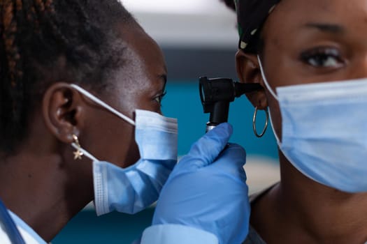 Closeup of african american otologist doctor with protective face mask against coronavirus checking patient ear using medical otoscope during clinical appointment in hospital office. Otoscopy tool