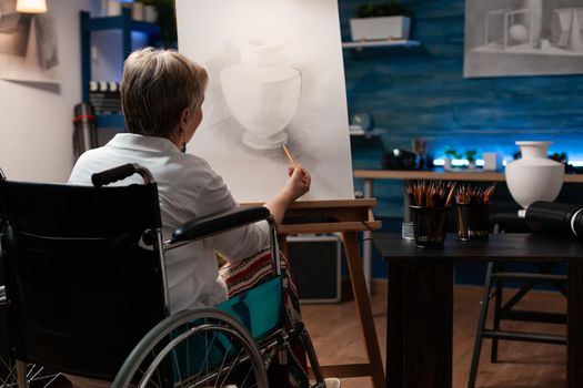Invalid aged artist drawing vase on canvas using pencil in artwork studio. Senior woman with handicap sitting in wheelchair while creating professional masterpiece for fine art