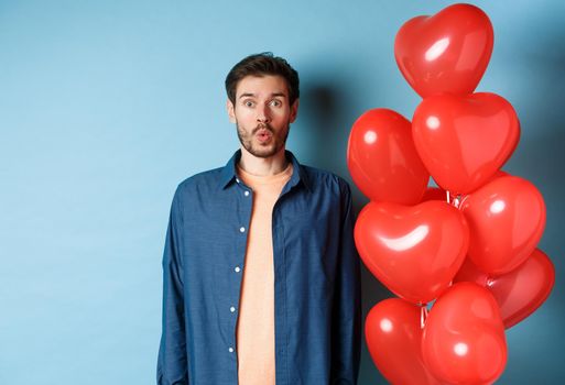 Happy valentines day. Man looking surprised and interested at camera, saying wow, standing near red hearts balloons, blue background.