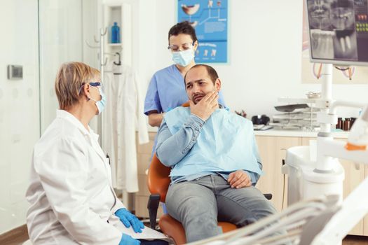 Sick patient complaining about toothache while talking with dentist before intervetion. Senior doctor speaking to man sitting on stomatological chair while nurse stomatology hospital office