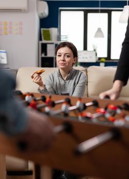 Woman eating pizza at foosball table after work having fun with soccer game at office. Worker enjoying drinks together with colleagues coworkers friends. Happy caucasian businesswoman