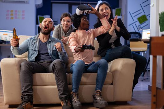 Diverse team of colleagues enjoy vr glasses experience after work at office while using controller joystick. Multi ethnic friends bonding and celebrating relaxation fun enjoyment