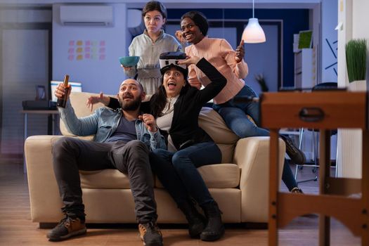 Team of multi ethnic coworkers using vr glasses after work for party at office. Group of diverse people enjoy gaming on console with controller joystick while having fun at celebration