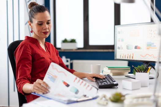 Business woman in start up company working on computer and analyzing charts on clipboard, sitting at desk. Successful concentrated manager working on deadline project typing.