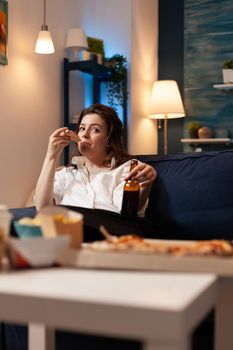Caucasian female relaxing on sofa eating pizza slice drinking beer during entertainment movie series unhealthy lunch night. Woman enjoying takeaway food home delivery. Fast-food pizzeria order