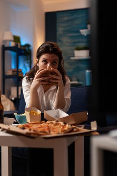 Portrait of woman holding delicious buger eating takeaway delivery food watching entertainment movie on television. Caucasian female sitting on couch tasting tasty junk-food enjoying night