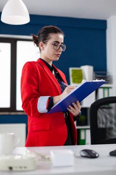 Entrepreneur standing in corporate office workplace taking notes on clipboard while doing financial analysis. Manager filling paperwwork checking charts on computer screen.