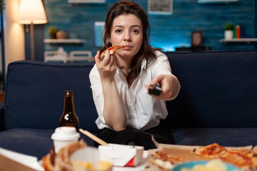 Portrait of caucasian female sitting on sofa eating slice of pizza enjoying takeaway food home delivered in living room. Woman relaxing in living room ordering tasty delicious meal lunch.