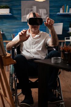 Invalid aged artist using vr glasses to draw masterpiece design on canvas and easel at fine art studio. Disabled senior man with modern headset sitting in wheelchair, for drawing hobby