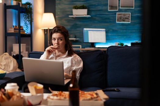 Woman eating tasty snack working on laptop computer while watching documentary movie series on television. Caucasian female enjoying takeaway food home delivered. Fastfood delicious order