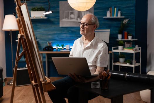 Senior artistic person using laptop for masterpiece drawing on canvas in workshop studio at home. Caucasian elderly artist with gadget creating fine art for professional artwork hobby