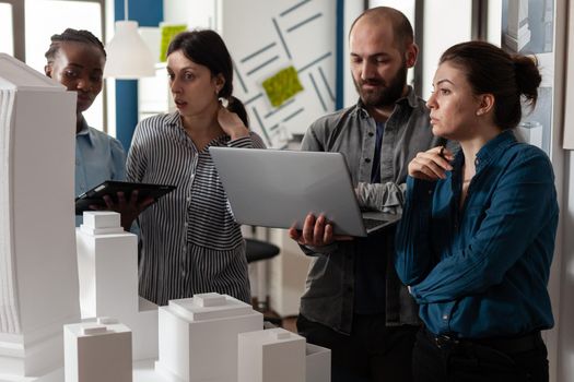 Architecture multi ethnic workers meeting at office analyzing blueprint plans on laptop computer tablet. Group working on maquette building model project design construction