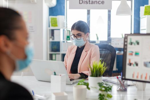 Woman entrepreneur working on laptop in new normal office with coworkers keeping social distnacing behind plastic shiled, wearing face mask as safety prevention during cornavirus flue outbreak.