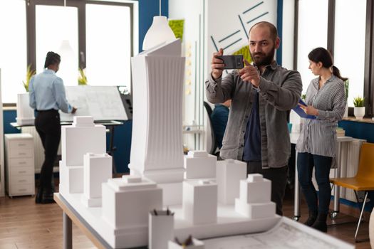 Architect profession man looking at maquette layout using smartphone to design building model plan maquette. Urban designer constructor working on professional modern construction