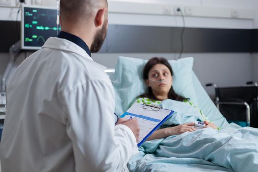 Oncologist practitioner doctor explaining sickness diagnose writing medical disease treatment on clipboard working in hospital ward. Patient sitting in bed recovering after medical surgery, health care system