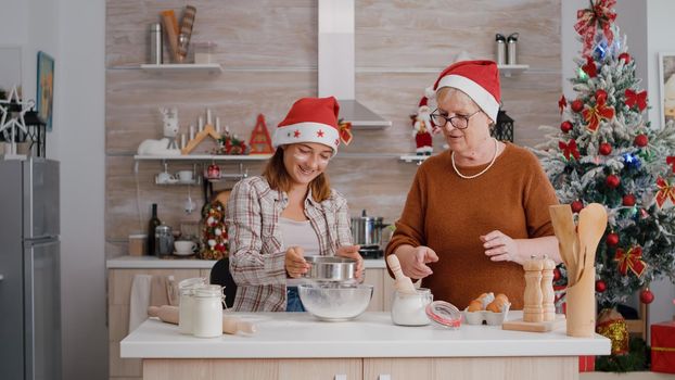 Grandmother explaining how to making traditional dessert dough to granddaughter preparing homemade delicious cookie in culinary decorated kitchen. Happy family enjoying christmas holidays