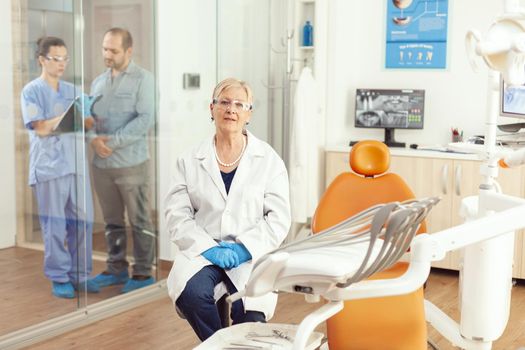 Portrait of smiling senior dentist woman in dental office while medical nurse talking with patient in background. Stomatologist doctor looking on webcam sitting on chair in stomatological clinic