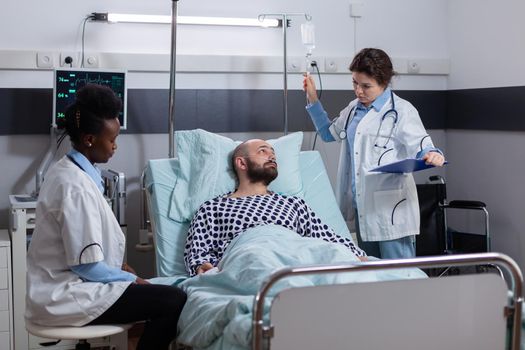 Physician doctor examining disease symptom writing medical treatment on clipboard in hospital ward. Afro-american nurse injecting fluids checking iv drip bag monitoring heart rate