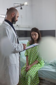 Patient discussing with doctor while sitting on bed during disease recovery in hospital ward. Practitioner writing sickness treatment on clipboard examining medical symptom