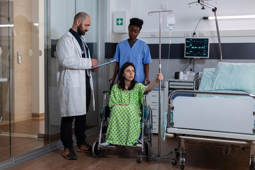 Therapist medic writing disease recovery on clipboard in hospital ward. Black woman asisstant putting sick patient in wheelchair for physiotherapy rehabilitation suffering leg accident