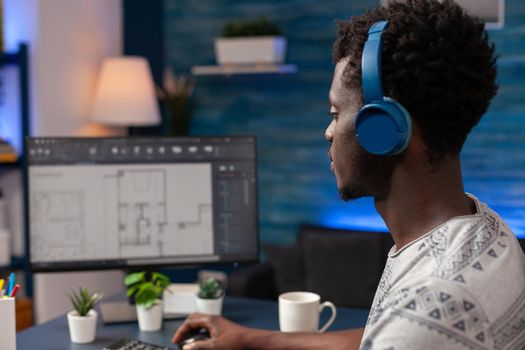 African american architect worker with headset sitting at desk in living room analyzing skyscraper construction plan. Young engineer working remote from home developing architecture model of building