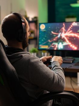 Cyber man with headset playing game in competition holding wireless controller in professional equiped studio. Excited player sitting on gaming chair looking at monitor for winning online championship