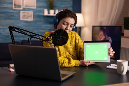 Influencer holding tablet with green screen and speaking into microphone during podcast in home studio. On-air production internet broadcast host streaming live content using chroma key mockup display.