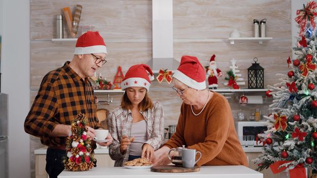 Family with santa hat speding christmas holiday together enjoying christmastime smiling in decorated kitchen. Grandparents drinking coffee while grandchild eating delicious chocolate baked cookies