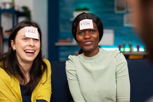 Multiracial friends enjoying friendship while playing guess who game attaching sticky notes on forehead. Mixed race people having fun, laughing together while sitting on sofa late at night.