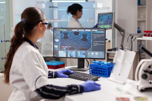 Biologist doctor checking DNA informations typing on pc in modern equipped laboratory. Scientists examining vaccine evolution in medical lab using high tech, chemistry tools for scientific research.