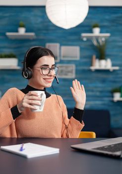 College student with glasses and cup of coffee in hand greeting her teacher during videocall meeting talking about university degree. Teenager with headphone on head sitting at desk table