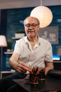 Senior caucasian man sitting at art studio desk with pencils for drawing hobby. Elderly artist with creativity looking at professional artistic tools and instruments on table in workshop