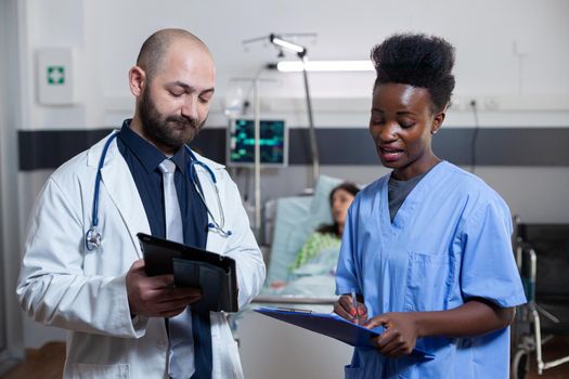 Man practitioner discussing with black assistant in hospital ward during medical consultation. Sick woman patient resting in bed while physician doctor monitoring illness disease