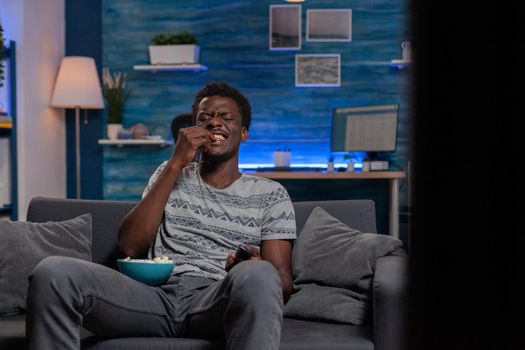 African american young adult laughing during comedy movie looking at television relaxing in living room. Black guy sitting on sofa while eating popcorn enjoying spending tine alone