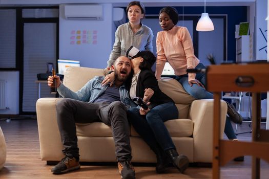 Multi ethnic team of mates play with vr glasses after work at office party. Diverse group of friends enjoy gaming experience with console controller joystick for victory celebration