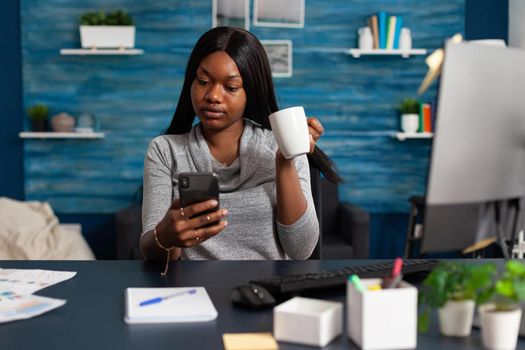 African american woman holding cup of coffee in hands socializing with friend using smartphone sitting at desk in living room. Black woman working remote from home browsing on social media