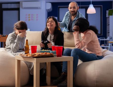 Diverse group of mates bonding while playing video game on tv console losing with joystick controller after work. Multi ethnic team enjoy office celebration party with snacks and drinks