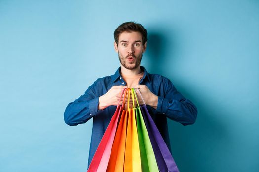 Excited shopper holding packages in paper bags and smiling amused, standing on blue background.