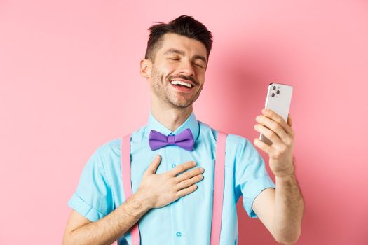 Technology concept. Happy caucasian man video chat and laugh at smartphone, standing over pink background.