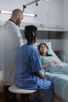 Sick woman patient resting in bed talking with medical doctor explaining disease symptom while writing treatment on clipboard in hospital ward. Black nurse monitoring adult during clinic appointment