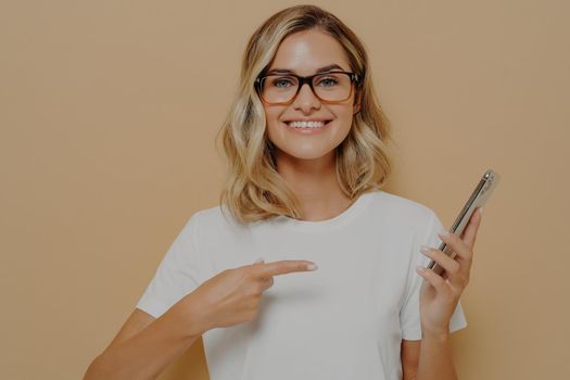 Portrait of impressed, excited young blond woman showing something awesome on display, pointing on mobile phone screen with forefinger and broadly smiling, brag with her recent win on casino app