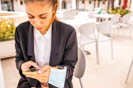 Young woman self entrepreneur texting on smartphone sitting at bar dehors with holding protective face mask in hand. Freelance girl taking breakfast at hotel cafe using technology. New normal habits