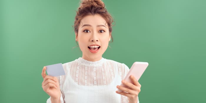 Happy young woman standing isolated over green background, holding mobile phone, showing plastic credit card