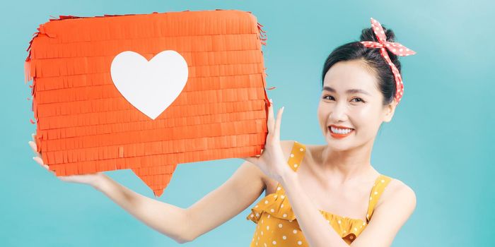 pretty smiling girl holding red paper with heart symbol and looking at camera isolated on blue