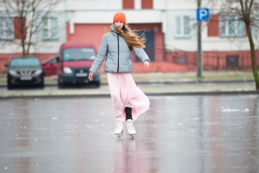 Girl teenager skates on ice in the city.Young girl in a long dress and a warm jacket on the ice.