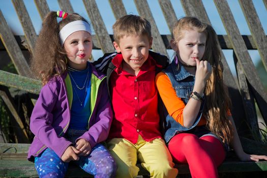 Funny little children: girls with bright makeup dressed in the style of the nineties and a boy in a red shirt are sitting on a bench by a skewed fence. Russian village children.