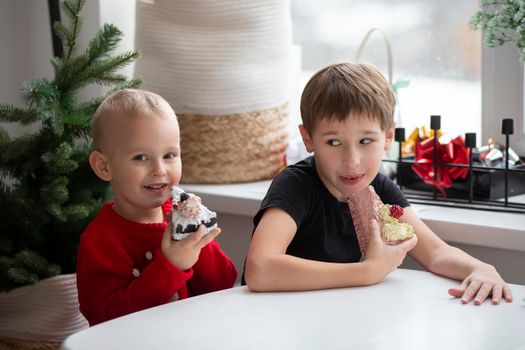 Children eat at Christmas. Two little boys with a piece of cake on the background of a festive Christmas tree.