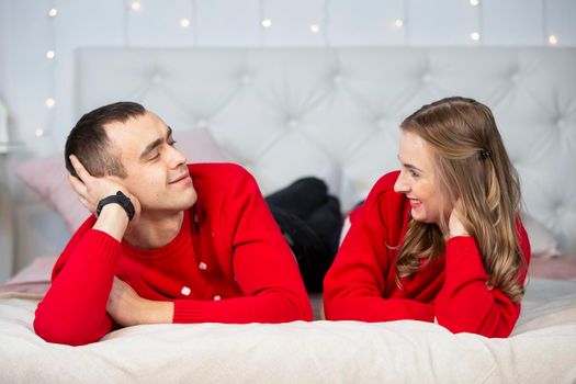 Handsome young man and woman in red sweaters lie on the bed and look at each other.