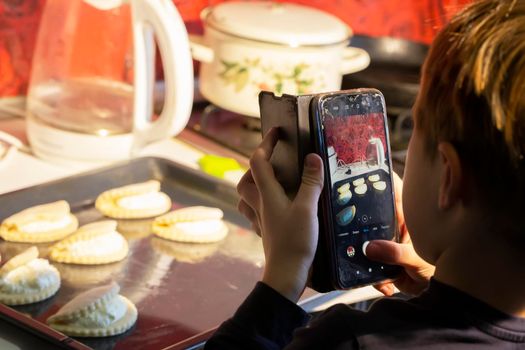 The child takes pictures of the cooking process on a smartphone. A boy with a mobile phone takes pictures of cookies on a pan.