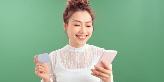 A beautiful portrait of a happy young girl showing plastic credit card while holding mobile phone isolated over green background